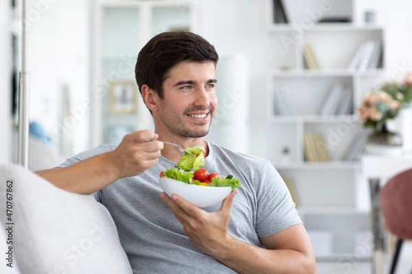 Fototapeta Handsome man eating fresh vegetable salad while sitting on the couch at home, healthy and vegan