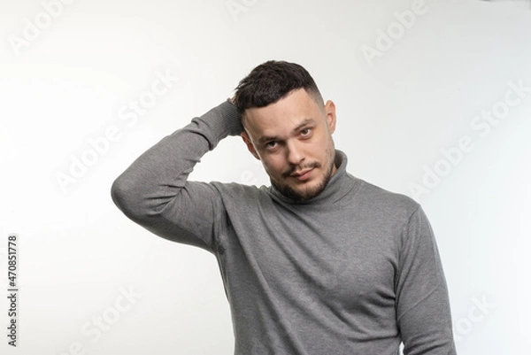 Fototapeta A young dark-haired man with a small beard looks in front of him with mockery and slight disappointment. Light background. One person, isolated.