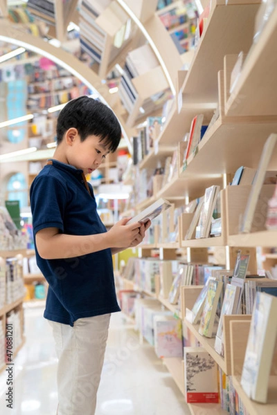 Fototapeta Young Asia boy standing in the modern and nice interior bookstore. Choosing and holding a book. Many bookshelf in the shop. Kid learning and education concept. Colorful book at library
