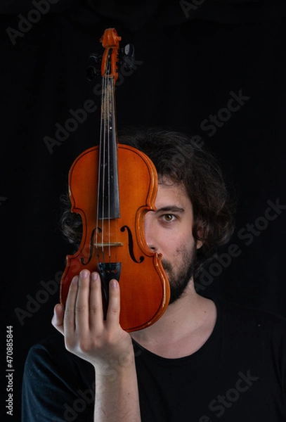 Fototapeta Portrait of a young dark-haired man with a beard, looking out from behind an old violin. Dark background. Selective focus.
