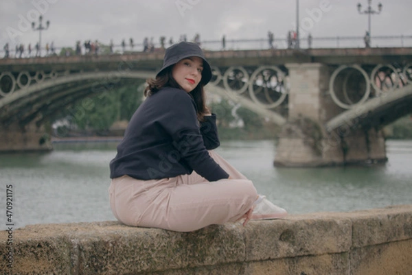 Fototapeta Caucasian girl sitting outdoors smiling at camera and Triana Bridge on the background. Travel during corona virus 2021-2022 concept.