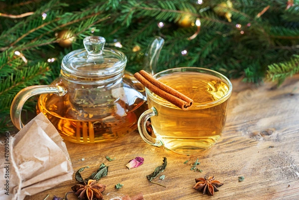 Obraz A glass teapot with herbal tea, a cup, a craft bag with herbs on a wooden table with a Christmas tree and garlands