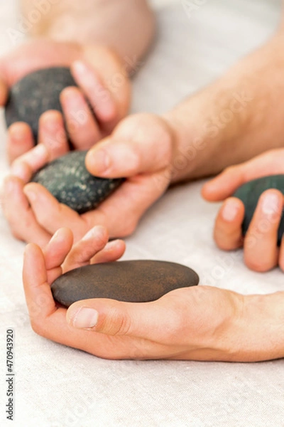 Fototapeta Four hands of male masseurs holding spa stones on white towel top view. Stone massage concept