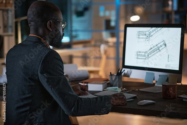 Fototapeta Rear view of African engineer sitting at the table in front of computer monitor and developing blueprint online during work at office