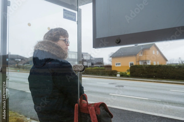 Fototapeta Man in blue coat with backpack is waiting for a bus. Modern glass bus stop on Norway. Cloudy day.