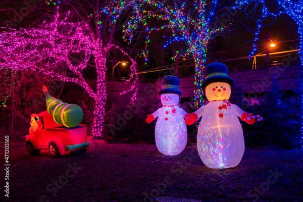 Fototapeta A pair of Christmas inflatable snowmen stand by as Santa drives a Christmas tree away in his truck.