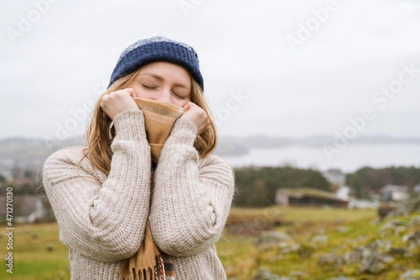 Fototapeta Young woman in knitted jaket holding scarf with hands over nose. Nature in background. Cloudy day in Norway. Closed eyes.