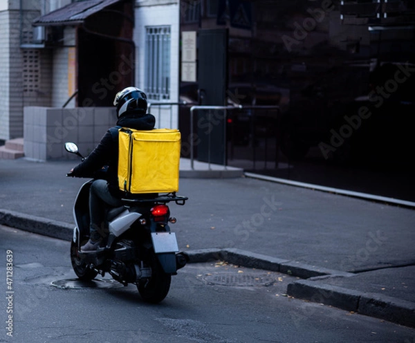 Obraz Delivery of food from cafes and restaurants, a courier on a scooter with a yellow backpack travels around the city. Fast food delivery to customers.