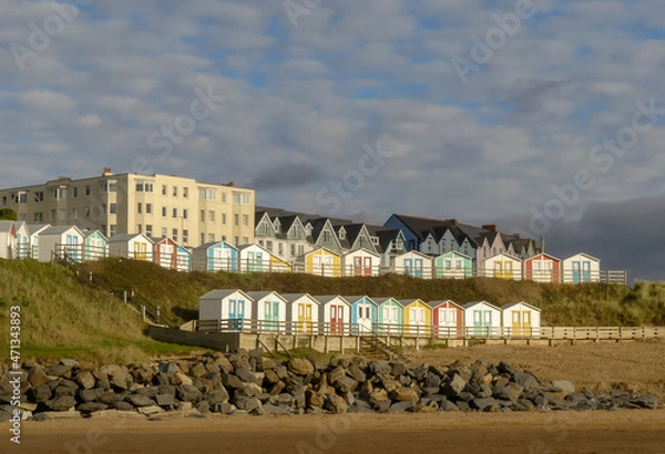 Fototapeta Rows of iconic colourful beach huts banked upon a hillside above a Cornish beach. Blue sky with dappled white clouds.  Outside landscape image with space for text. Bude, England.