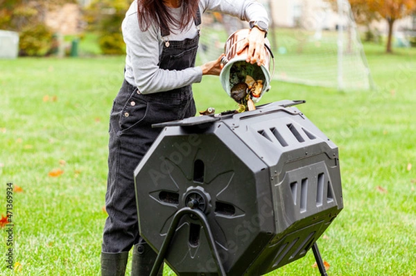 Fototapeta A woman is dumping a small bin of kitchen scraps into an outdoor tumbling composter in backyard garden. These plastic units with metal legs can turn around for better aeration and quick composting.