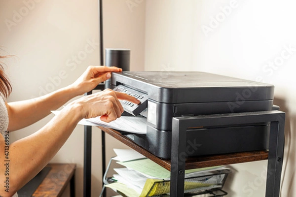 Fototapeta Concept image for office work. A woman is placing or removing paper. There is a wireless photocopier, scanner, printer combo device on top of a shelving unit at a small office setting.