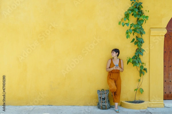 Fototapeta front view of woman standing with smartphone isolated on yellow facade. Horizontal view of latin american woman sightseeing on yellow wall using technology. People and travel concept.