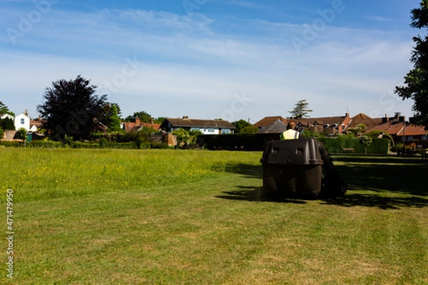 Fototapeta A council worker riding on a sit on lawnmower cutting the grass in a public park on a hot summers day