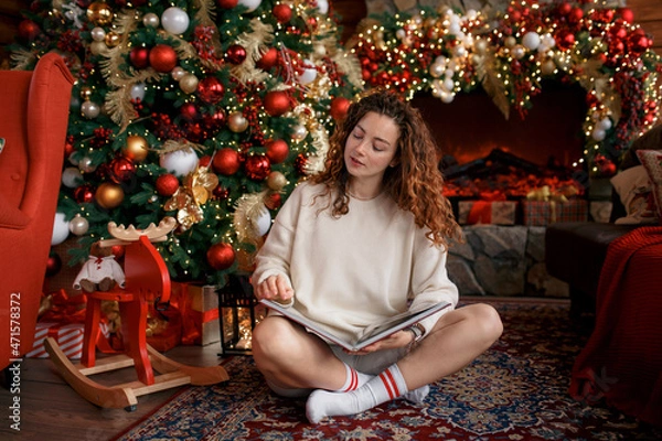 Obraz A beautiful girl is sitting on a sofa in a Christmas interior. Christmas tree, decorated fireplace, lots of gifts, Christmas theme. Cozy atmosphere. red-haired girl reading a book.