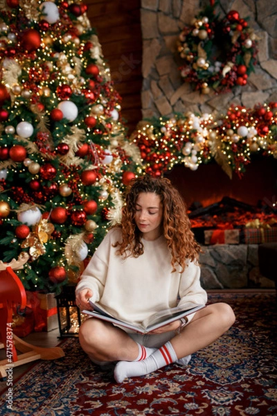 Obraz A beautiful girl is sitting on a sofa in a Christmas interior. Christmas tree, decorated fireplace, lots of gifts, Christmas theme. Cozy atmosphere. red-haired girl reading a book.