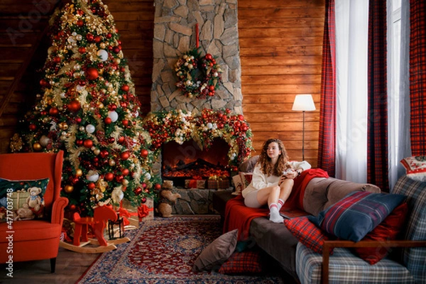 Obraz A beautiful girl is sitting on a sofa in a Christmas interior. Christmas tree, decorated fireplace, lots of gifts, Christmas theme. Cozy atmosphere. red-haired girl reading a book.