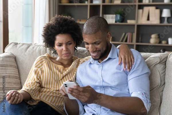 Fototapeta Happy mixed race guy using smartphone, confused suspicious girlfriend looking at boyfriend phone screen. Couple using online app on cellphone, chatting on internet, shopping, making virtual order