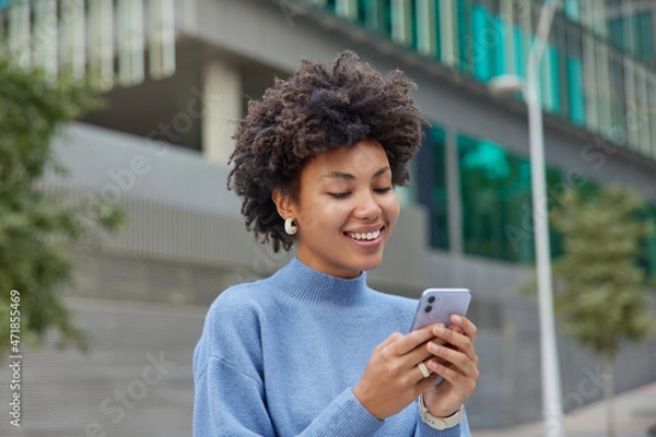 Fototapeta Half length shot of positive glad woman in blue turtleneck poses in urbanity with mobile phone explores new city downloads application has chat conversation. People technology and free time.