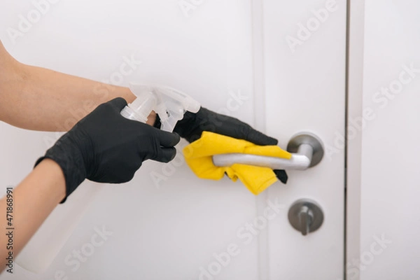 Fototapeta Cleaning door handle with yellow wipe in black gloves and sanitizer spray. Disinfection in hospital and public spaces against corona virus. Woman hand using towel for cleaning