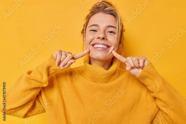 Fototapeta Portrait of cheerful young woman smiles broadly indicates index fingers at face wears casual jumper expresses positive emotions isolated over vivid yellow background. Look at my brilliant smile