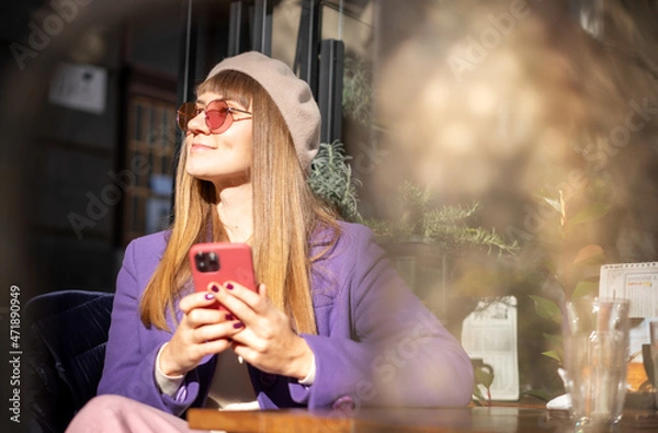 Fototapeta Stylish happy young woman with a smartphone in her hands while sitting in a street cafe in the city