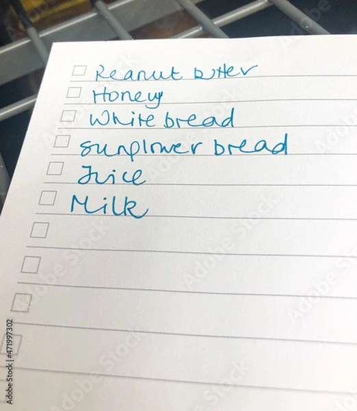 Fototapeta London, England - January 22 2021 - A shopping list on lined paper including peanut butter, honey, white bread, sunflower bread, juice and milk.  Image features intentional blur and has copy space.