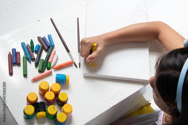 Obraz cute girl with light blue bow on her head, painting a blank canvas with yellow crayons, drawing the sun, photo view from above.