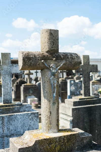 Fototapeta Stone cross with Jesus and graves with more crosses in the cemetery blurred behind