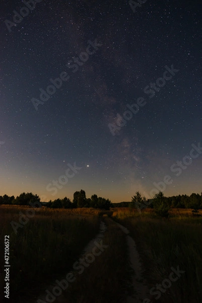 Fototapeta A large panorama, a country road stretching away into the fields to the forest. Starry sky with the Milky Way overhead