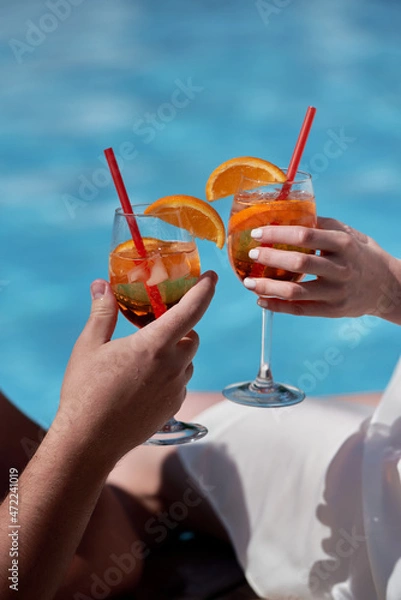 Obraz Couple hands toasting with aperol spritz cocktails on summer party by the pool, copy space. Event celebration concept. Traditioanal italian aperitif.