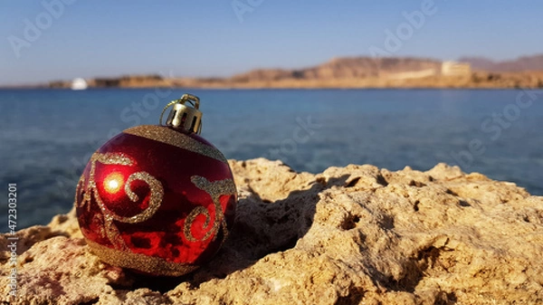 Obraz Christmas tree decoration in the form of a red ball on the background of the sea and the beach. Christmas in warm countries