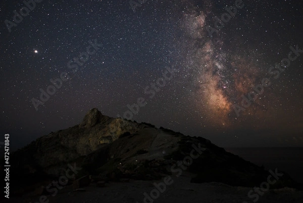 Fototapeta A large panorama of a beautiful view of the sea among the mountains and a beautiful starry sky with the Milky Way. The city by the sea glows at night in the distance