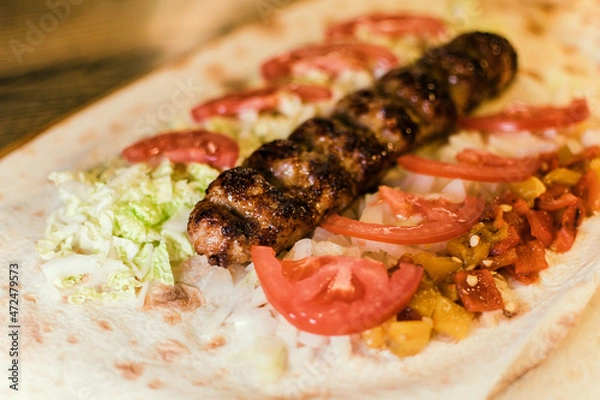 Fototapeta Shawarma with meat kebabs, with sauce, tomatoes, peppers, cheese, herbs, cabbage, onions and garlic on ruddy pita bread, lying on a wooden board