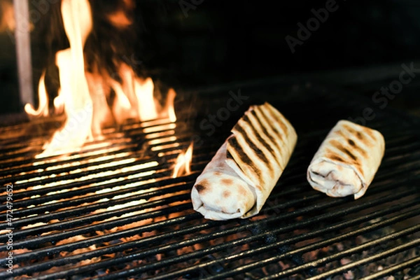 Fototapeta Shawarma with meat kebabs, on charcoal, on a fire, on a grill wire rack, with sauce, tomatoes, pepper, cheese, herbs, cabbage, onion and garlic in a ruddy pita bread, dark background