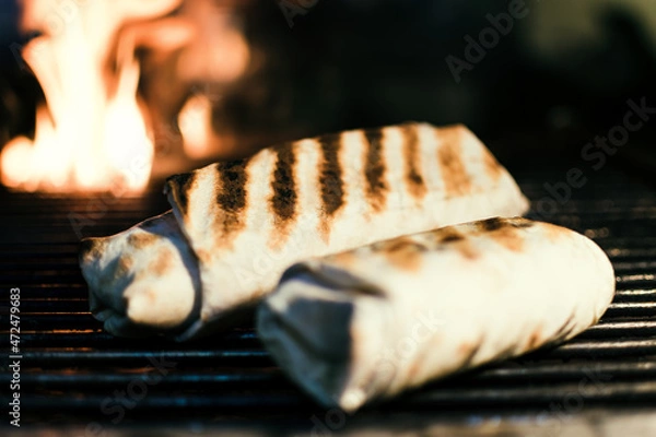 Fototapeta Shawarma with meat kebabs, on charcoal, on a fire, on a grill wire rack, with sauce, tomatoes, pepper, cheese, herbs, cabbage, onion and garlic in a ruddy pita bread, dark background