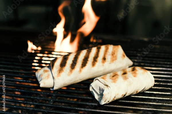 Fototapeta Shawarma with meat kebabs, on charcoal, on a fire, on a grill wire rack, with sauce, tomatoes, pepper, cheese, herbs, cabbage, onion and garlic in a ruddy pita bread, dark background