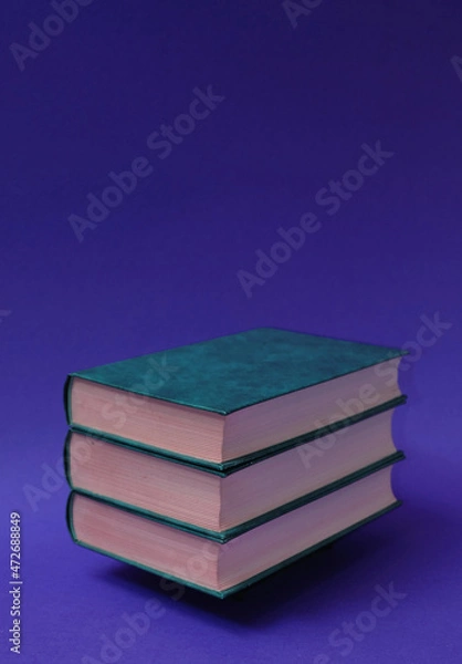 Fototapeta Three books, hardcover, stacked horizontally, one on top of the other, on a blue-violet background. Text space. Reading, leisure concept. Minimal style.