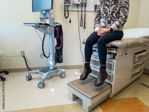 Fototapeta Inside of an  exam room with a bed, some medical devices, a computer. A woman is anxiously waiting for the doctor. Generic image for health care industry, disease, patient and doctor.