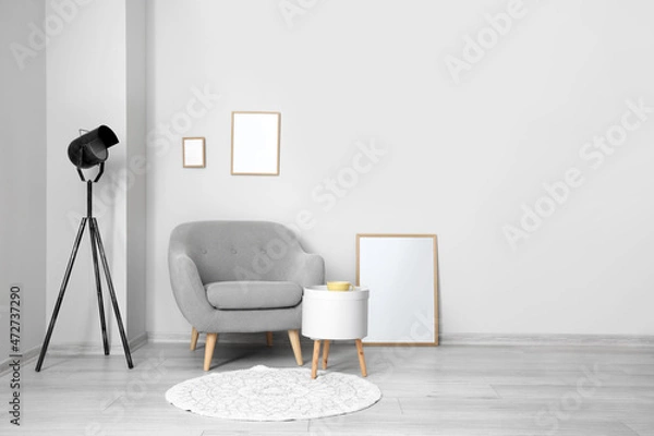 Fototapeta Blank frames, grey armchair with table and lamp near light wall