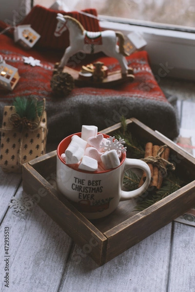 Fototapeta A mug with marshmallows is on the table in a wooden box, next to a sweater and a toy horse.