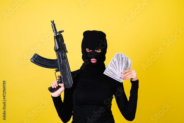 Fototapeta Young sexy beautiful woman with bright pink lips, balaclava on her head, holding a fan of 100 dollars and weapon machine gun, hacker, robbery, fraudster