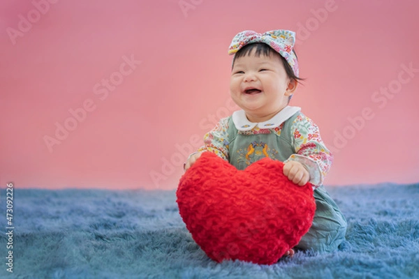 Fototapeta Asian baby valentine with red heart smiling and sitting on pink background. Cute 6 months baby sitting with copy space use as concept of valentine, love, baby or kid department in hospital.