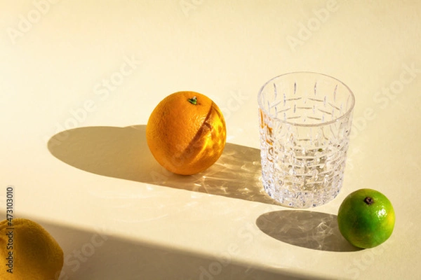 Fototapeta empty crystal glass under hard light, orange's, lime's and lemon's oval shadows, glitter reflection on beige background, food art composition
