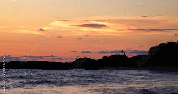 Fototapeta Silhouetted people on the rocky beach in the paradise island of Sri Lanka. The spectacular evening sky and the calm ocean waves create a romantic evening vibe.
