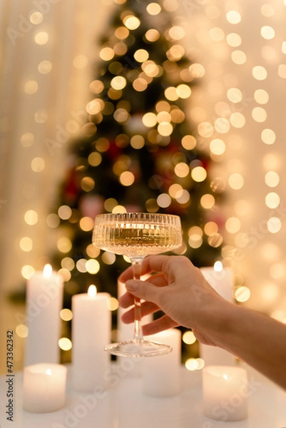 Fototapeta A woman's hand raises a glass of champagne. Table with candles, Christmas tree in lights in bokeh
