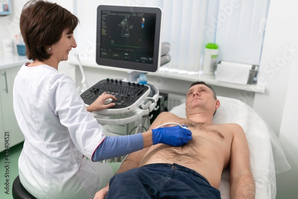 Fototapeta Woman doctor makes heart check of patient man using modern equipment in a cardiology clinic. Female cardiologist is screaning patient heart with ultrasonography, looking at screen. Healthcare concept