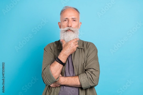 Fototapeta Photo of think old man touch beard wearing khaki shirt isolated over blue color background