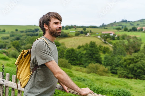 Fototapeta Panoramic view of caucasian man with backpack in green mountain. Horizontal view of backpacker traveling outdoors with travel destination in the background. People and travel concept.
