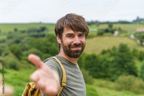 Fototapeta Front view of caucasian man reaching the hand to the camera. Horizontal view of backpacker traveling outdoors with travel destination in the background. People and travel concept.