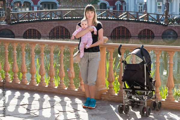 Fototapeta Mom with a baby girl and a stroller on the background of a pond and a bridge